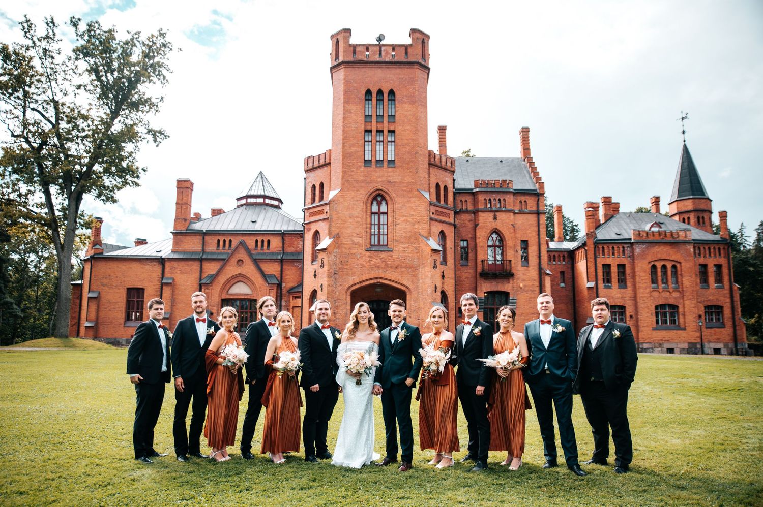 Wedding party posed in front of a grand red brick castle-like building with towers.
