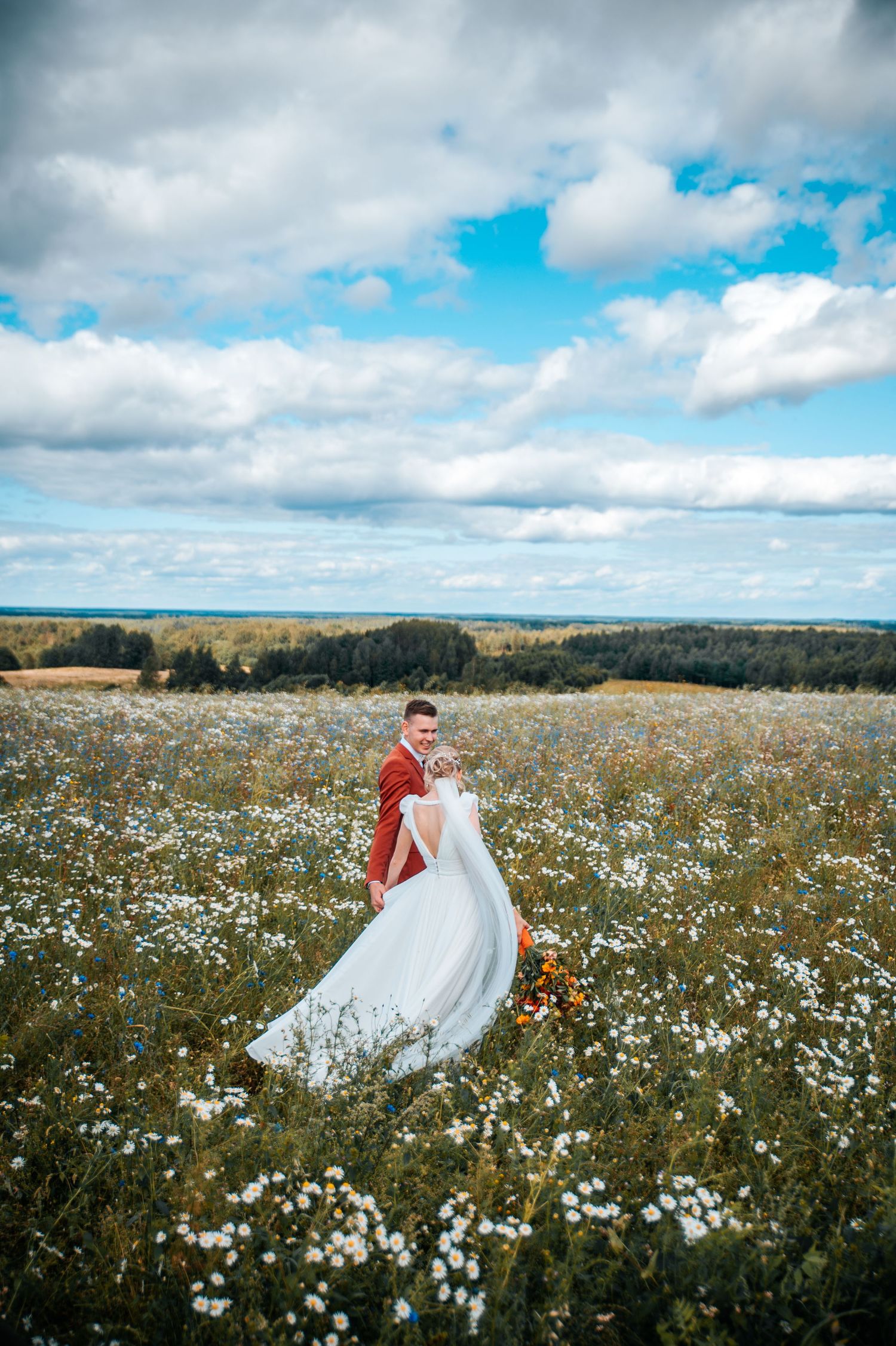 A couple dances in a picturesque field of white wildflowers under a cloudy blue sky.