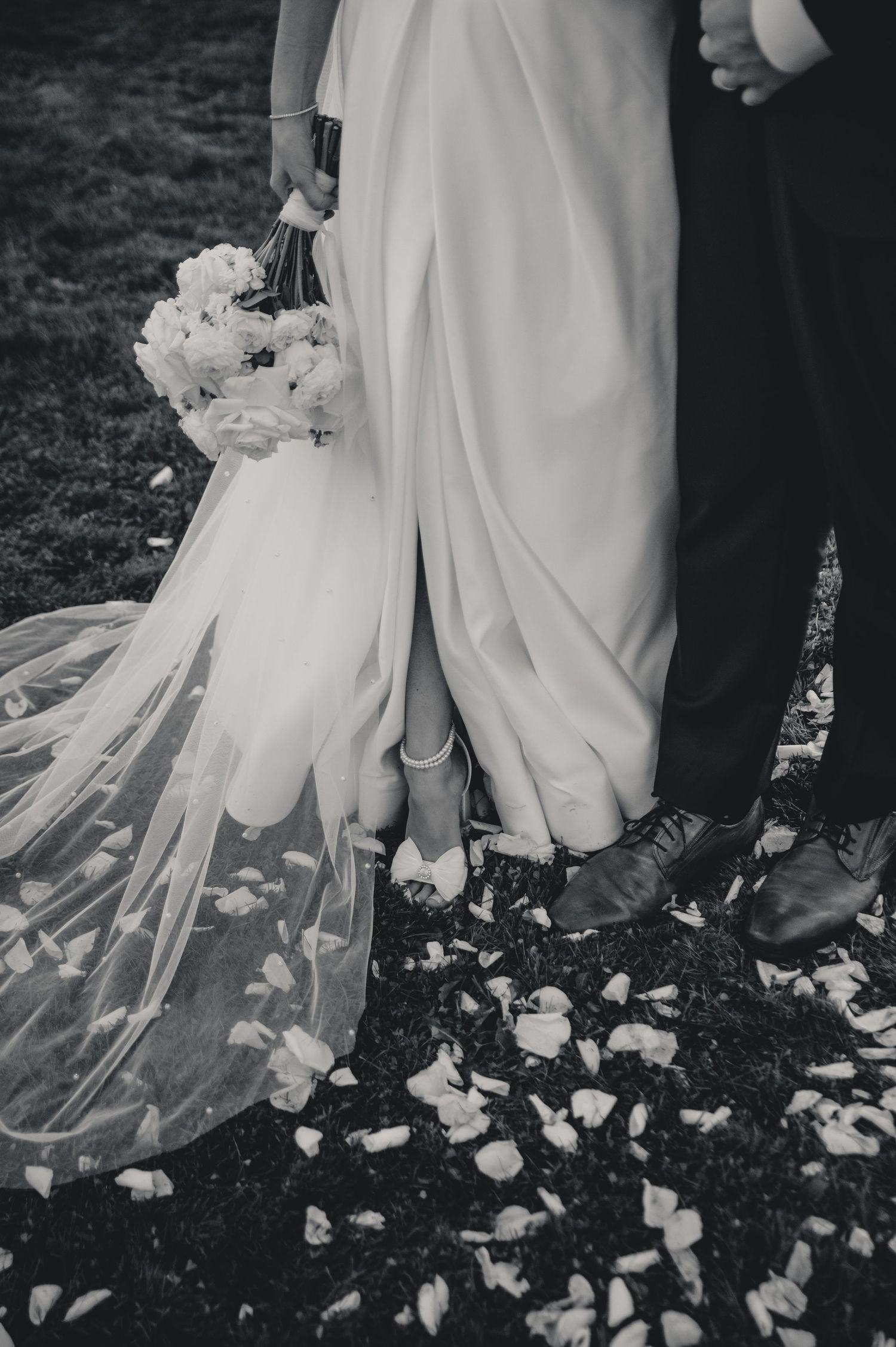 Black and white wedding photo series showing rose petals scattered around the hem of a flowing white bridal gown.