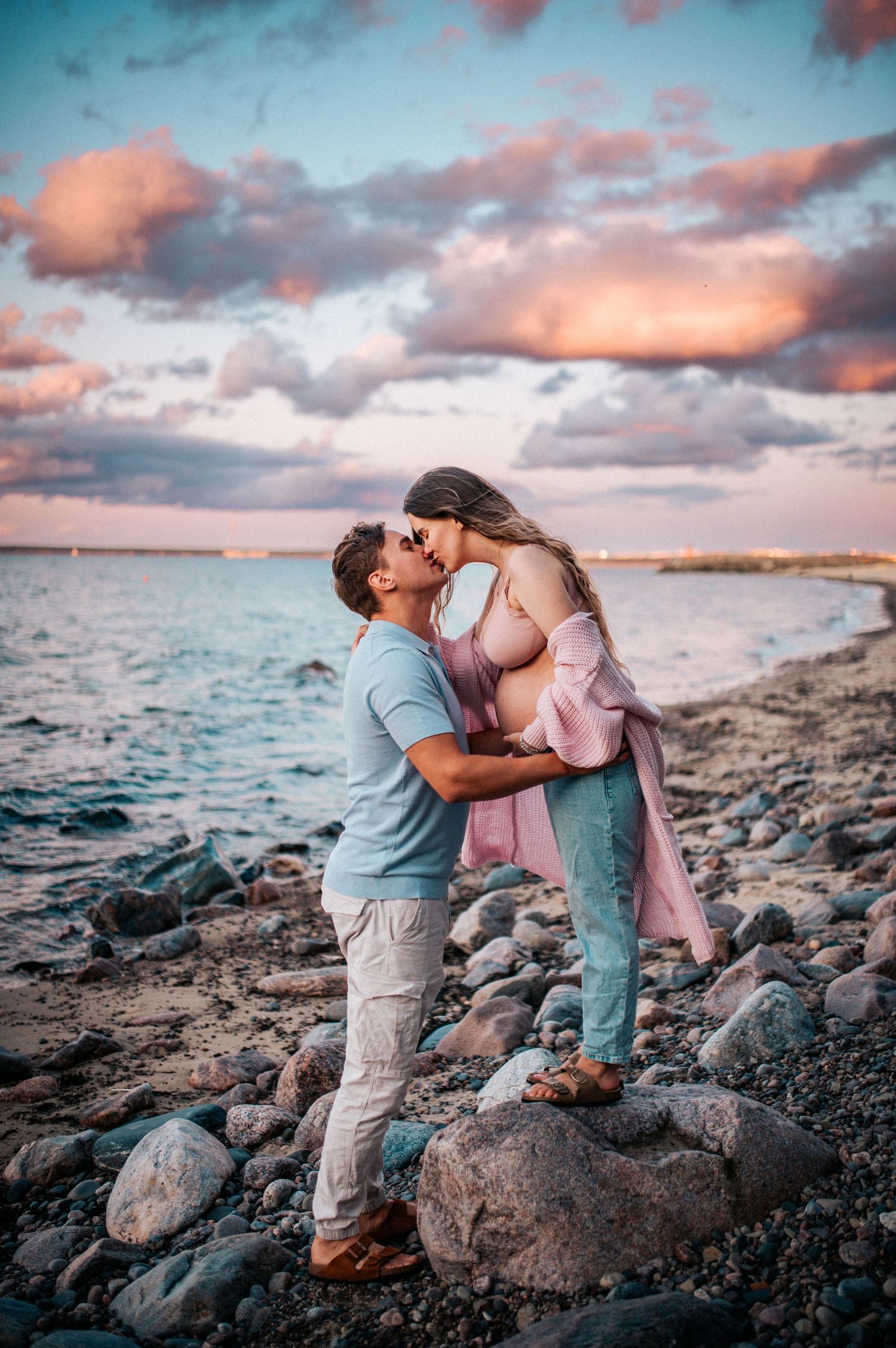 A romantic sunset photoshoot on a rocky beach with turquoise water and pink clouds in the background.