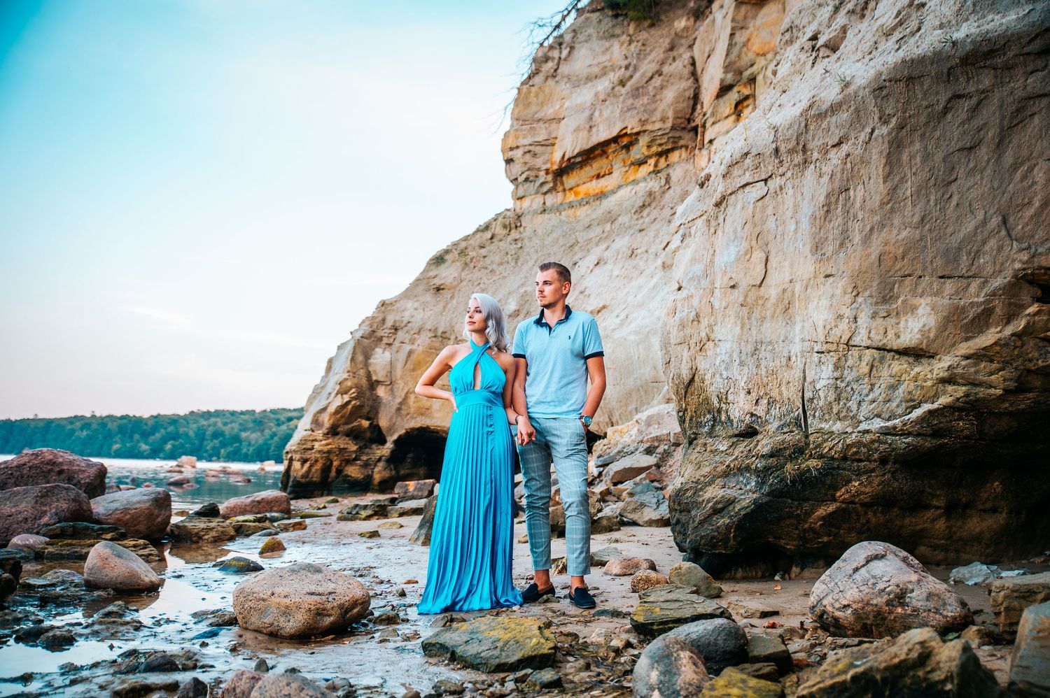 A couple stands together on rocky coastal terrain beneath a towering limestone cliff during a seaside photoshoot.