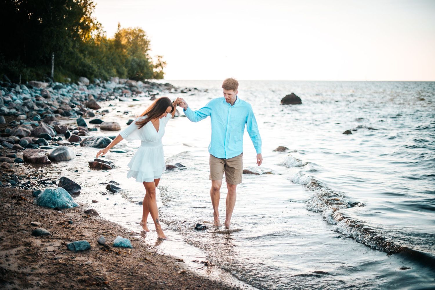 A couple walks along a rocky beach shoreline during sunset, creating a romantic coastal atmosphere.