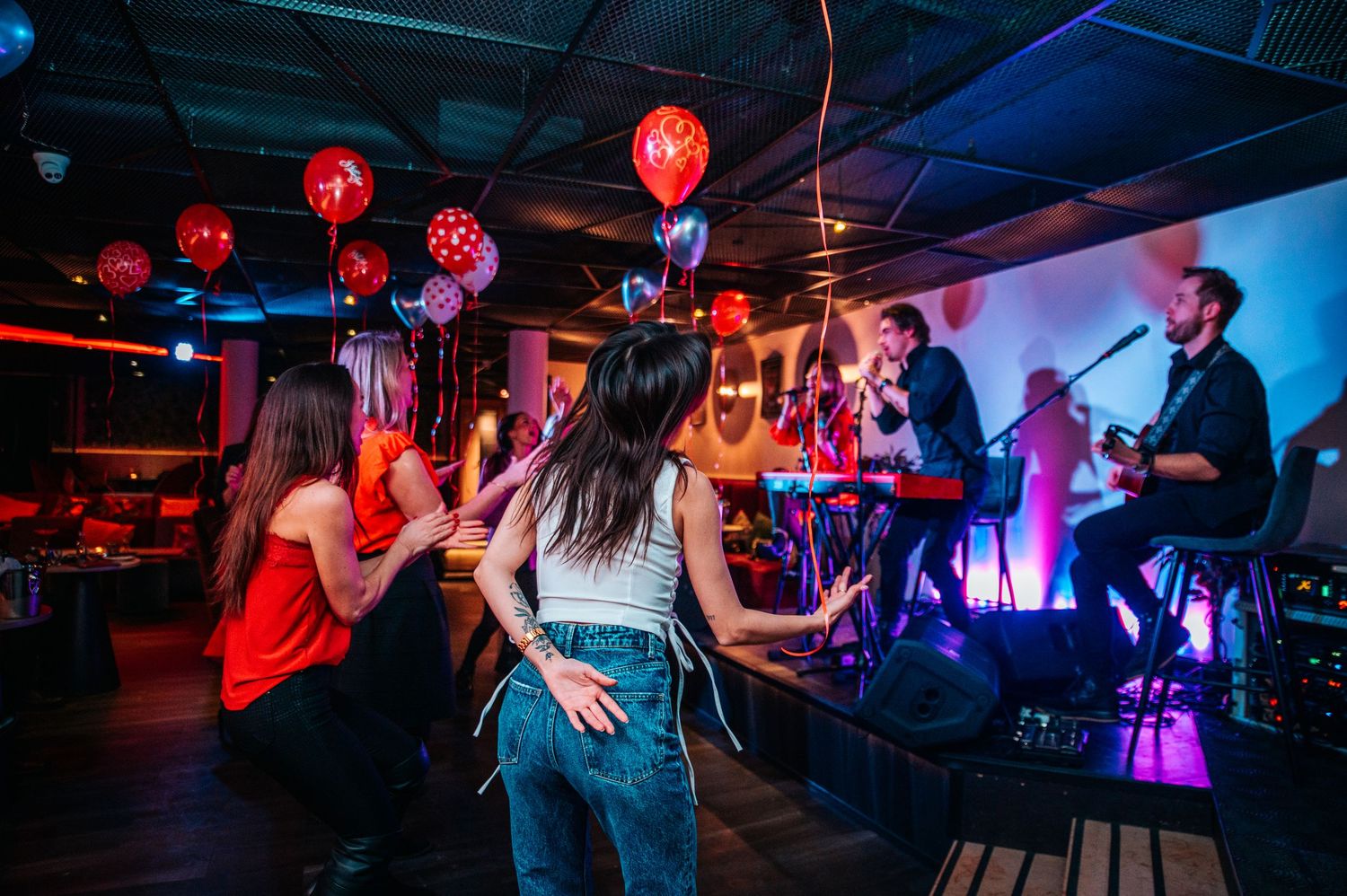 Nightclub scene with red lighting and crowd dancing on dark dance floor.