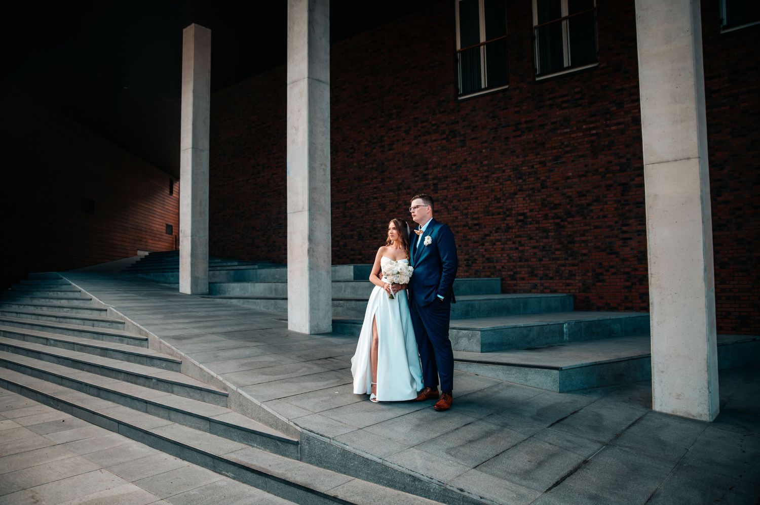 A couple in wedding attire stands in the shadows of a modern brick building's columns and steps.