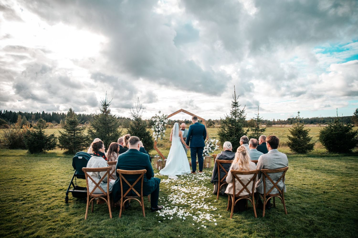 Intimate outdoor wedding ceremony on a grassy hillside with wooden chairs and dramatic cloudy sky overhead.
