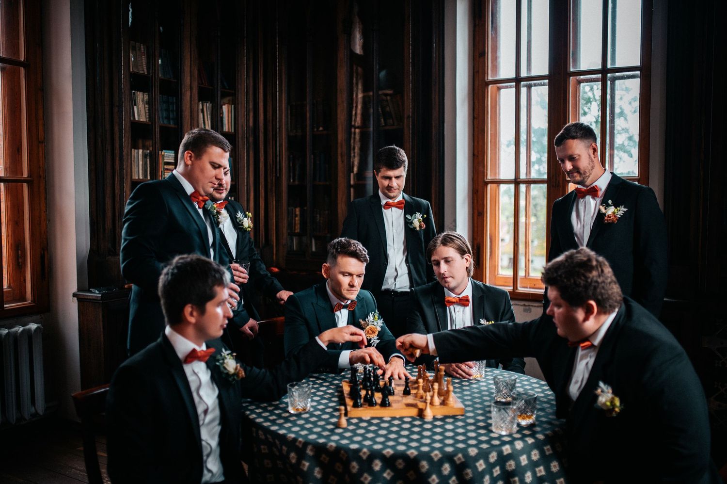Groomsmen in black tuxedos gather around a checkered table for drinks and laughter in an elegant dark wood room.