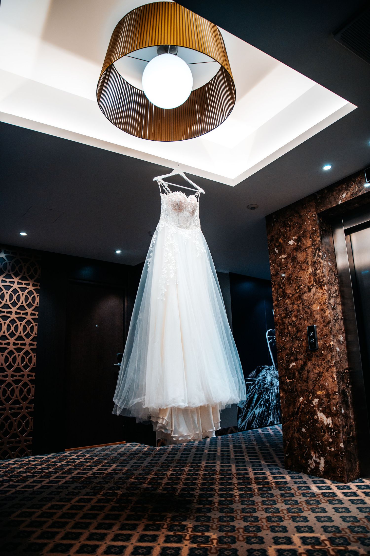A white tulle wedding dress hangs elegantly from a modern light fixture in a dark interior.