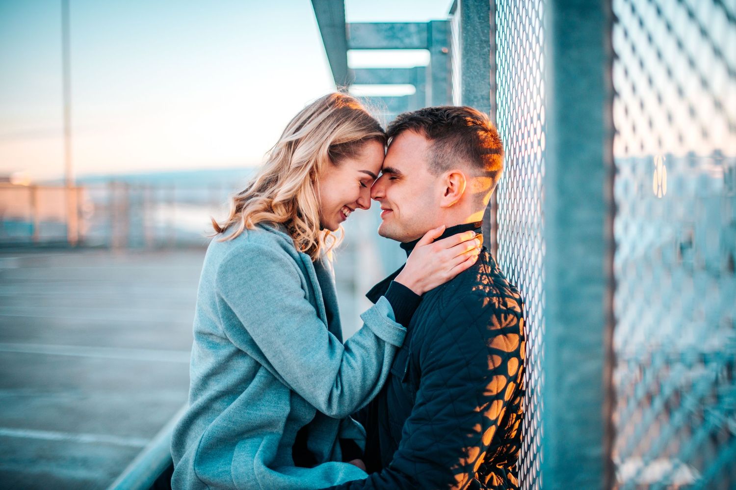 A romantic couple embraces in front of an industrial backdrop with soft lighting and chain link fencing.