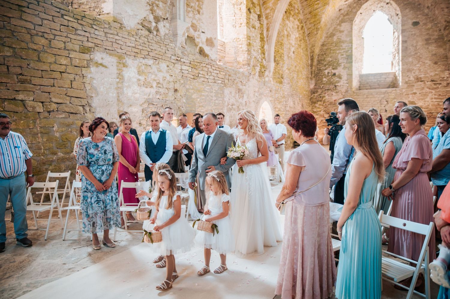 Wedding guests gather inside a rustic stone church with arched windows during a ceremony.
