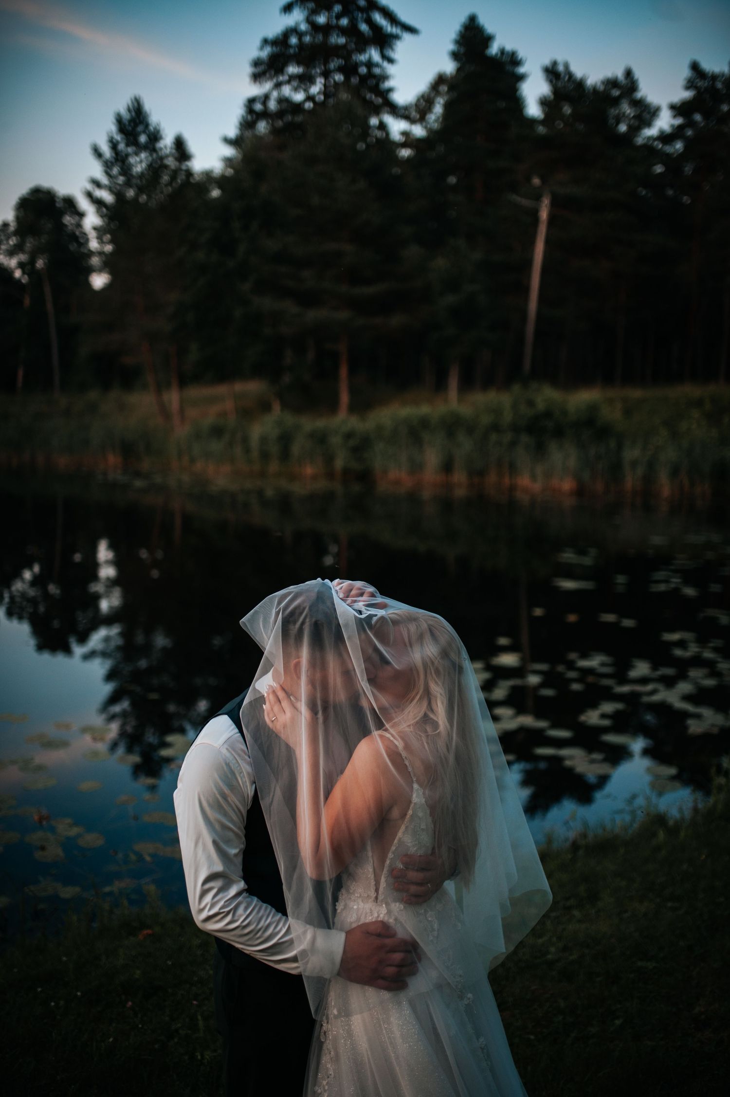 Romantic silhouettes of a couple embracing under a veil by a serene lake at dusk with pine trees in the background.