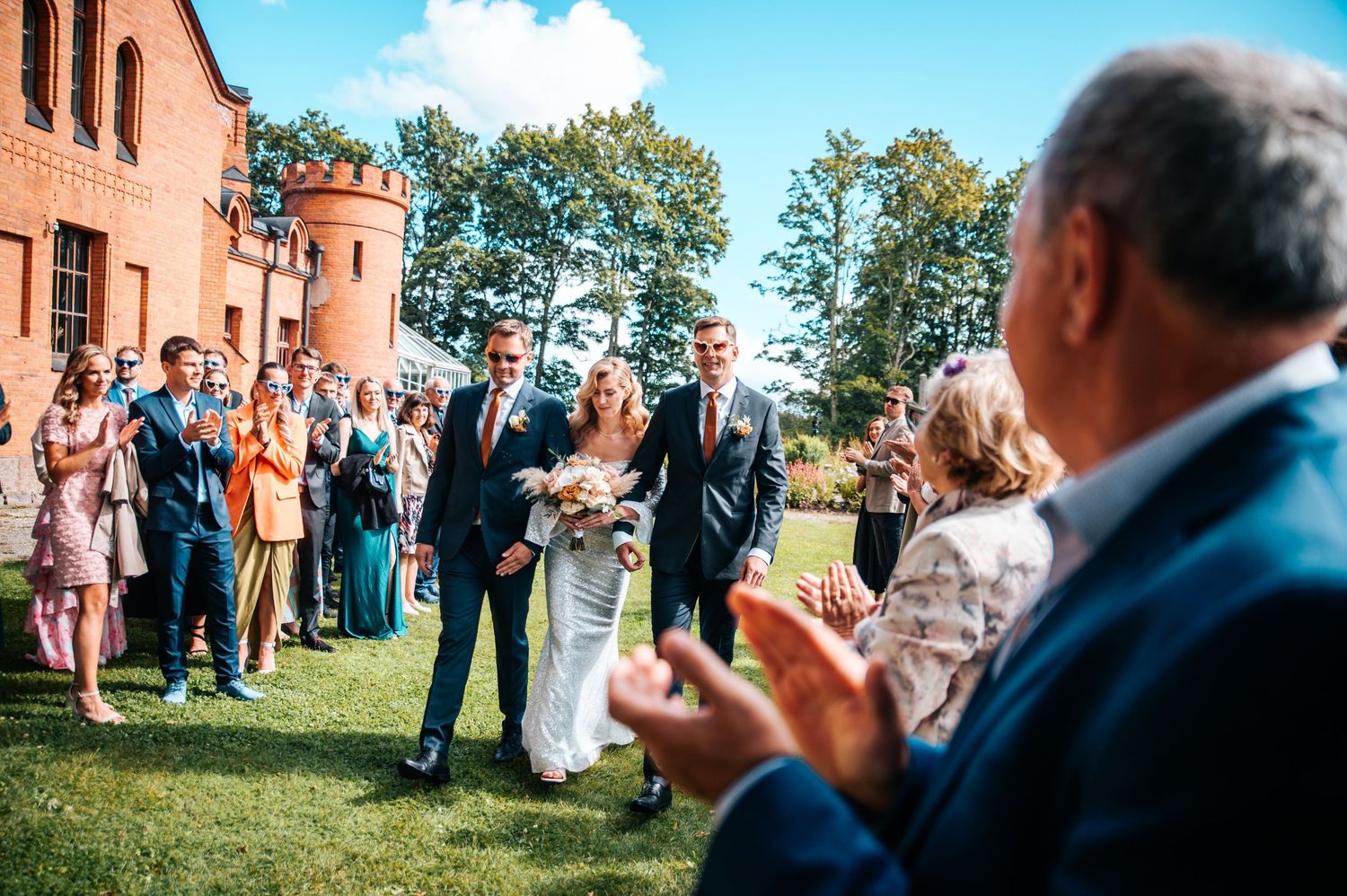 Wedding guests gather outdoors on a grassy lawn during a sunny celebration with brick buildings in the background.