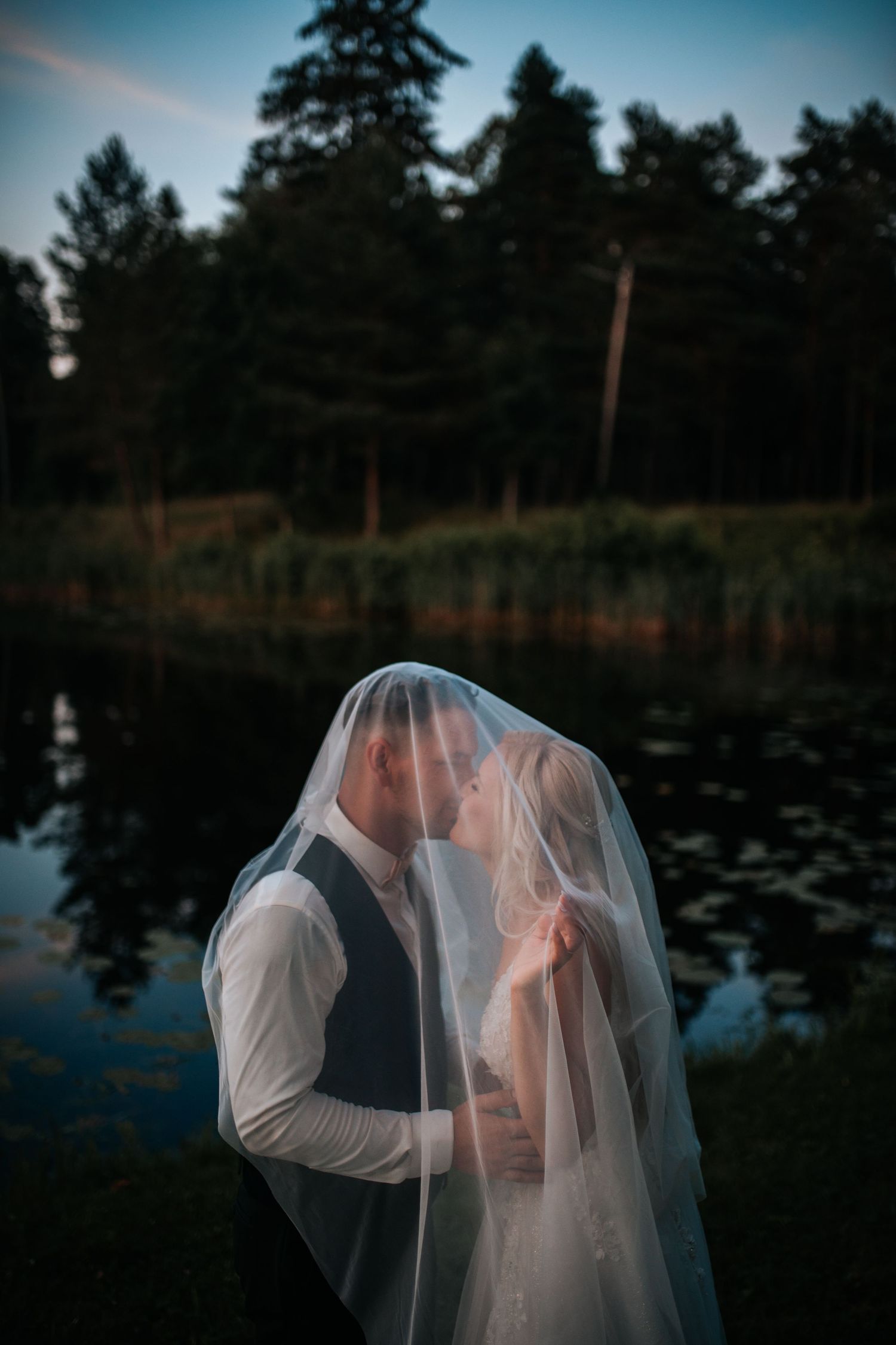 A romantic couple embraces under a flowing veil against a dark pond and forest backdrop at dusk.