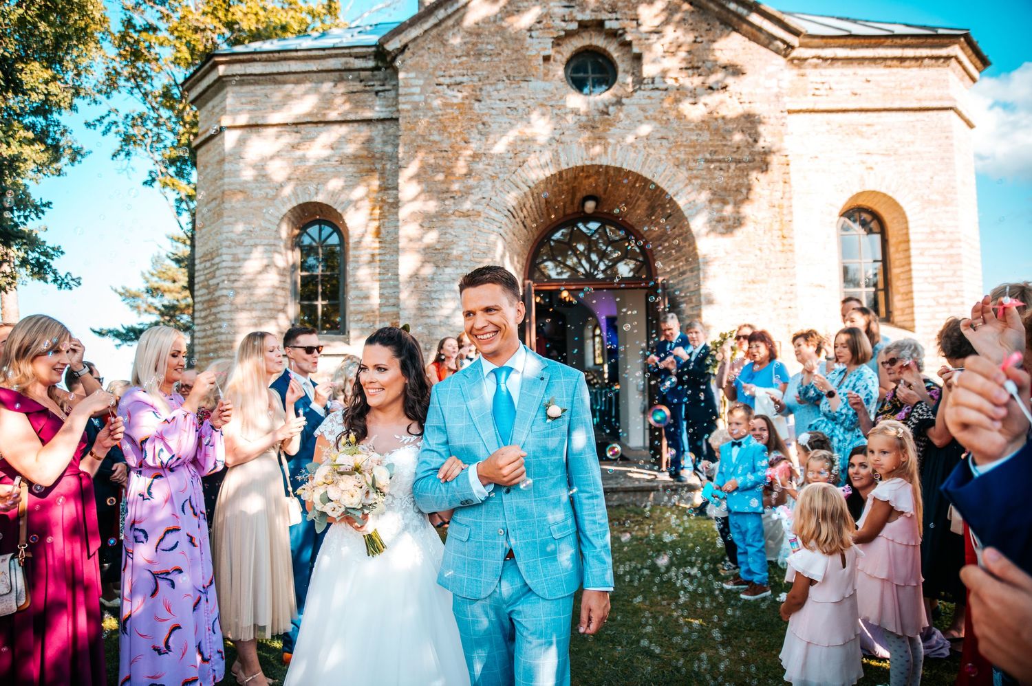 Wedding guests celebrate with bubbles outside a historic stone church on a sunny autumn day.