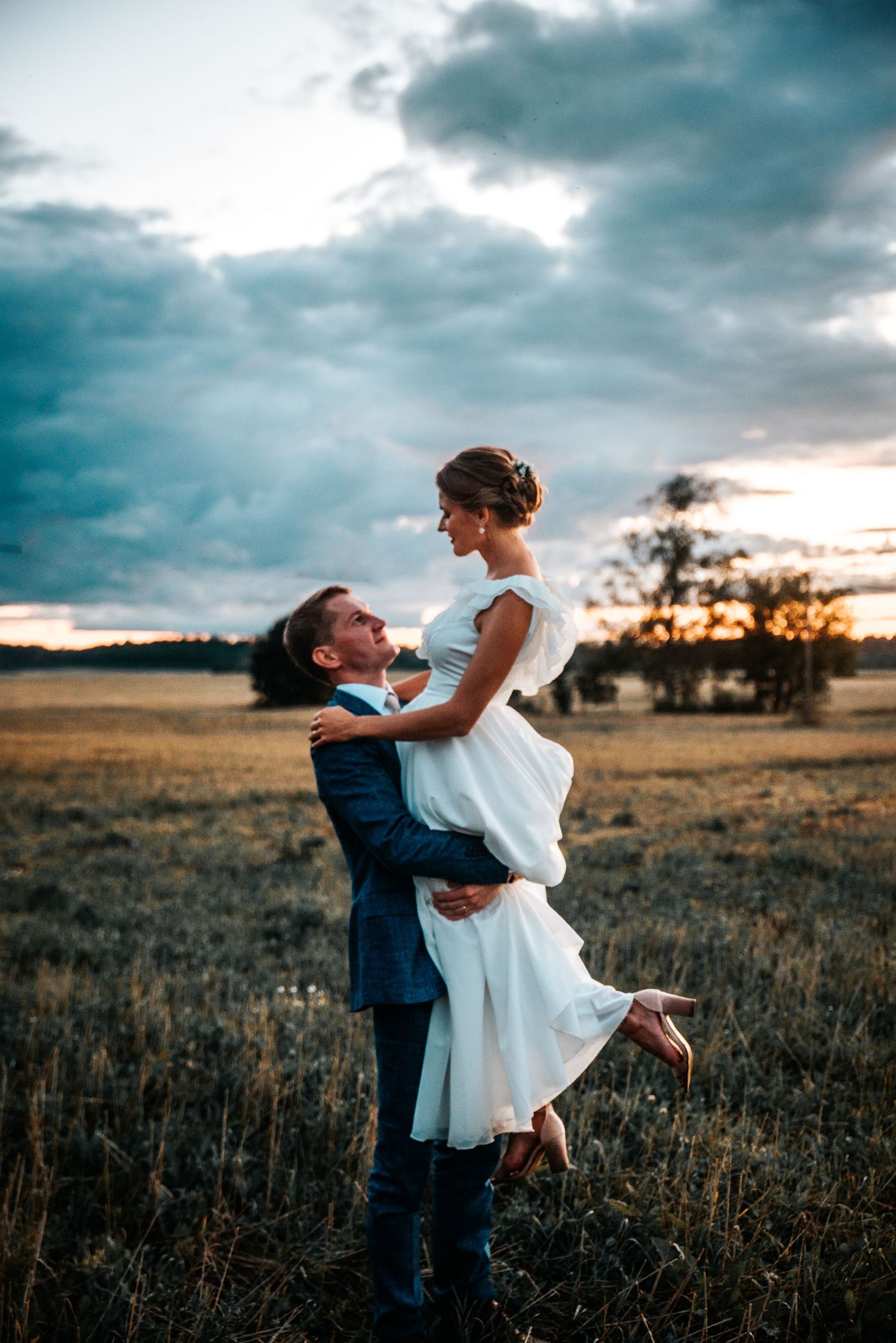 Romantic sunset wedding portrait of couple embracing in a golden field with dramatic cloudy sky.
