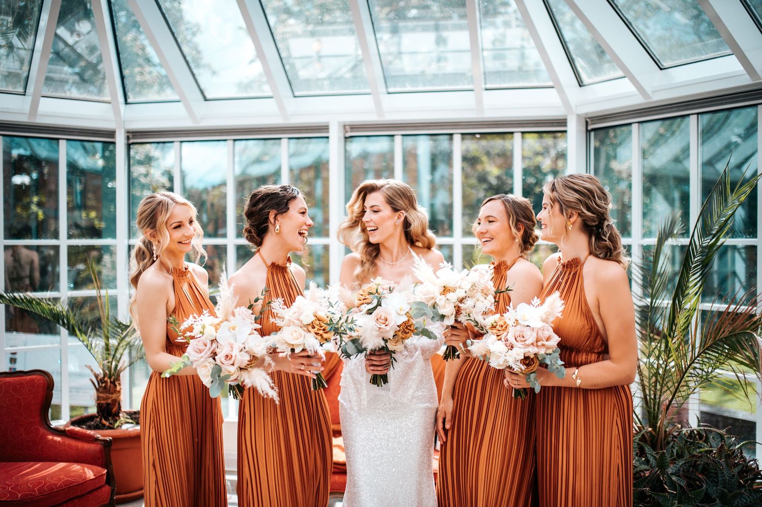 Bridesmaids in rust-colored dresses share joyful moments during an elegant greenhouse wedding celebration.