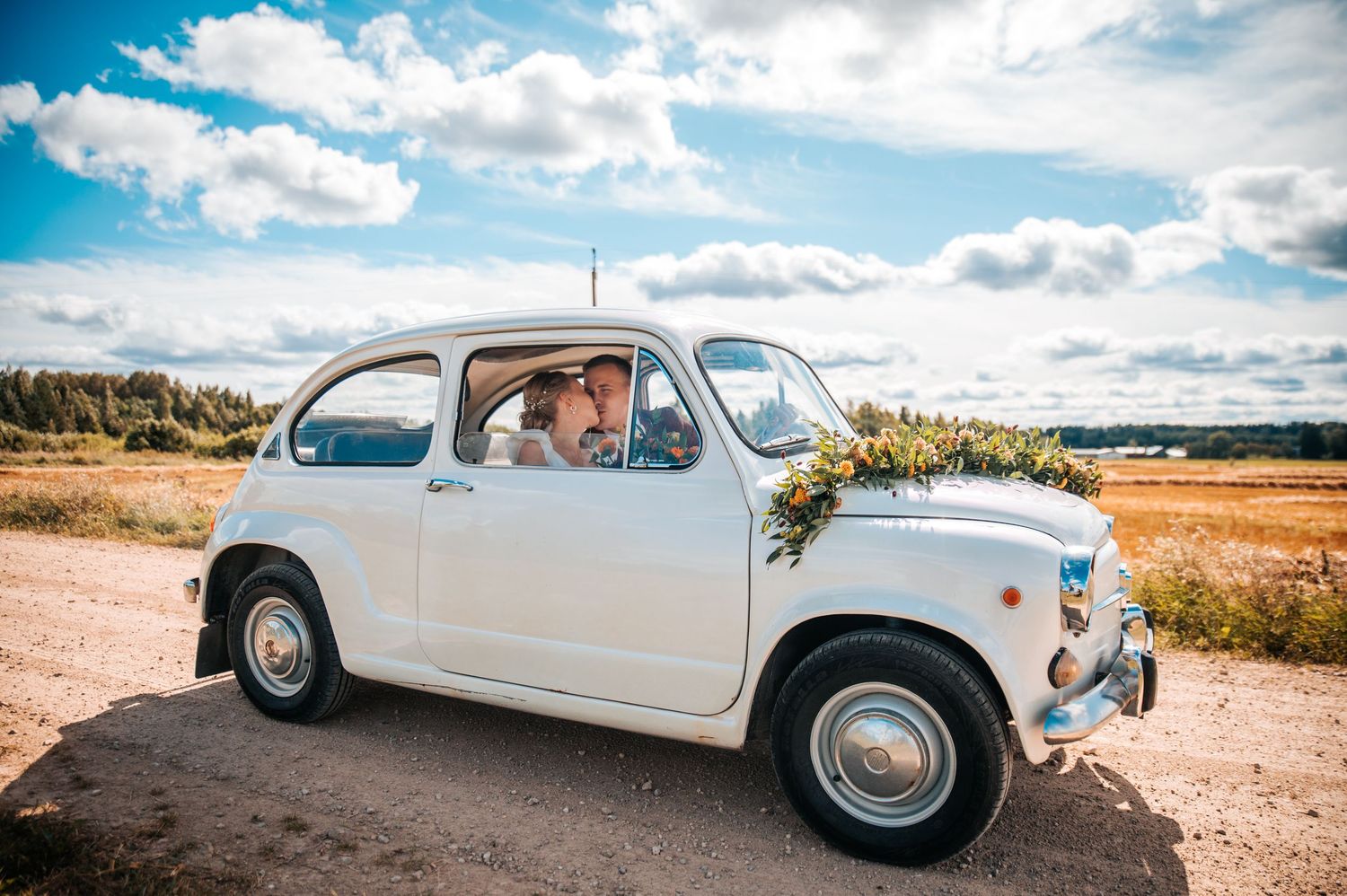 A vintage white Volkswagen Karmann Ghia car parked on a dirt road against a scenic blue sky with white clouds.