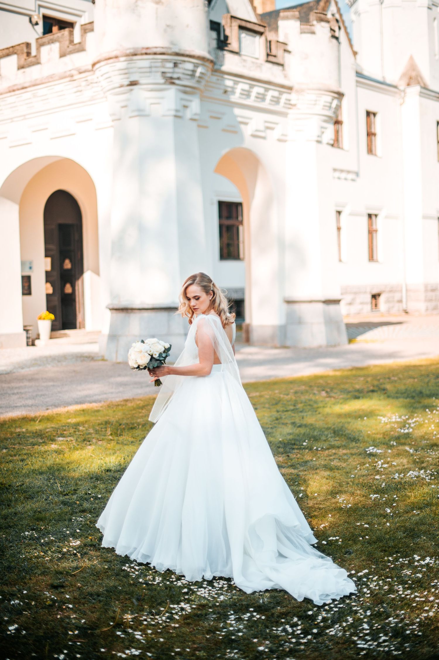 Bride in flowing white dress poses gracefully on grass near historic white church building.