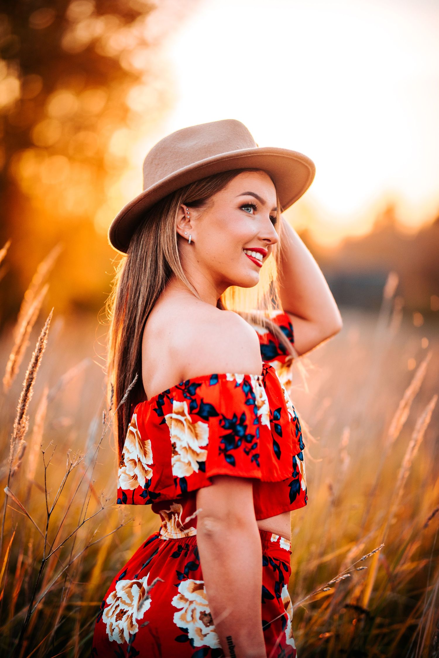 Woman in red floral dress and hat poses in golden sunset light among tall grass.