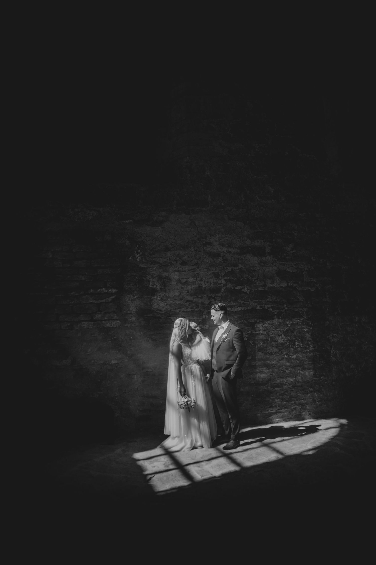 A dramatic black and white wedding photo shows a couple standing in a beam of light against a dark stone wall.