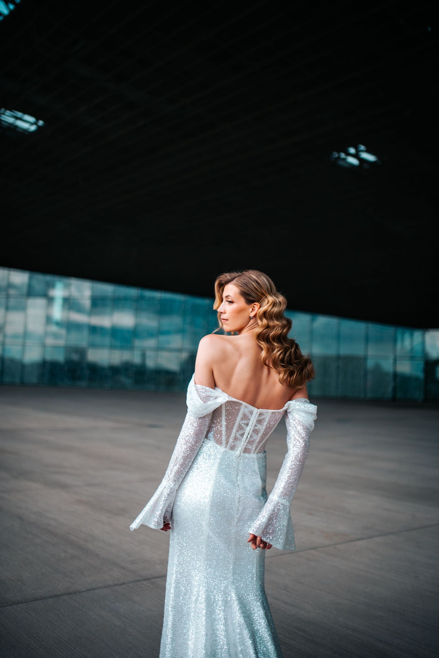 Bride in off-shoulder lace wedding gown with bell sleeves poses against modern glass building.