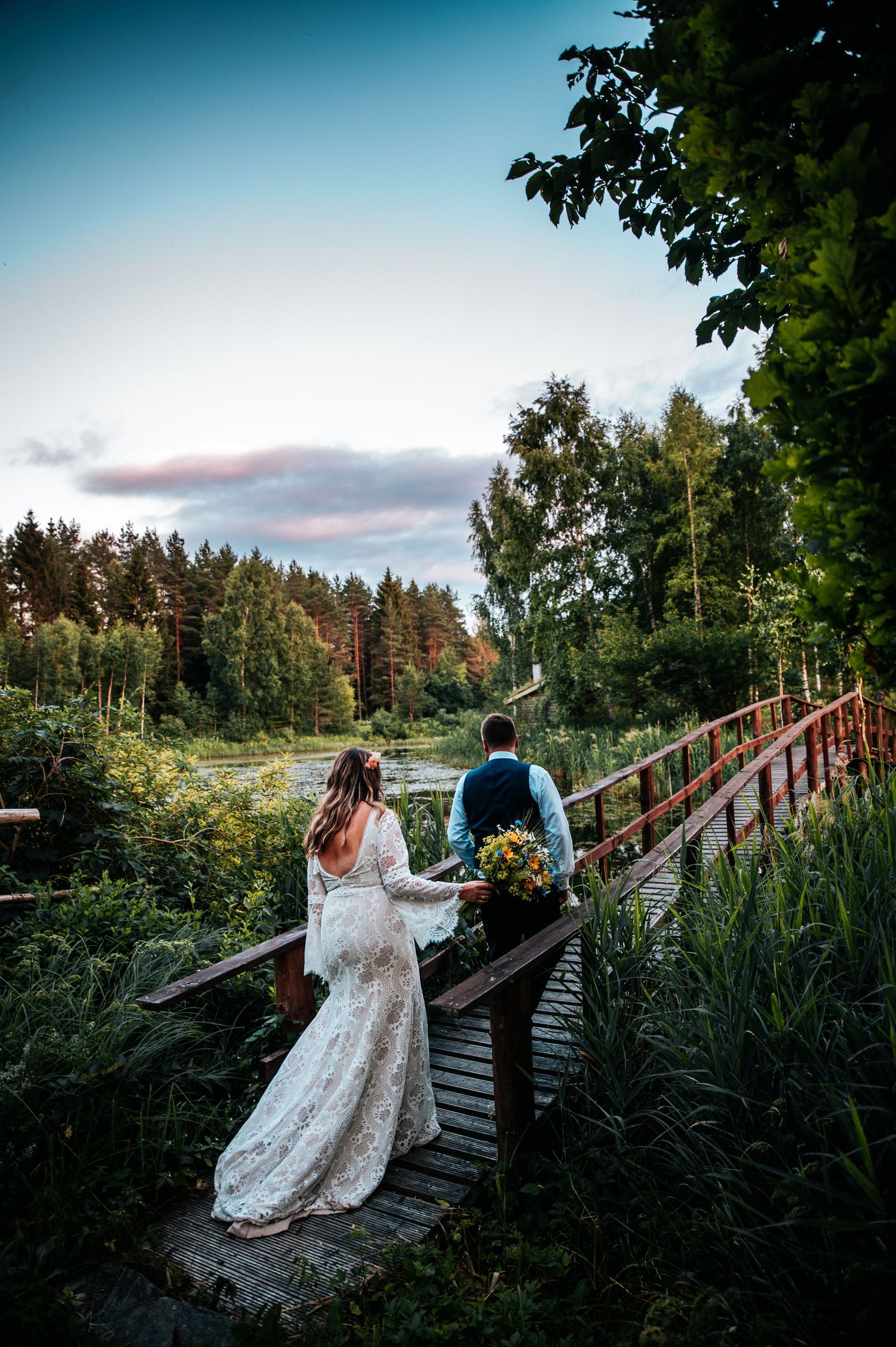 A wedding couple walks across a rustic wooden bridge surrounded by lush greenery at dusk.
