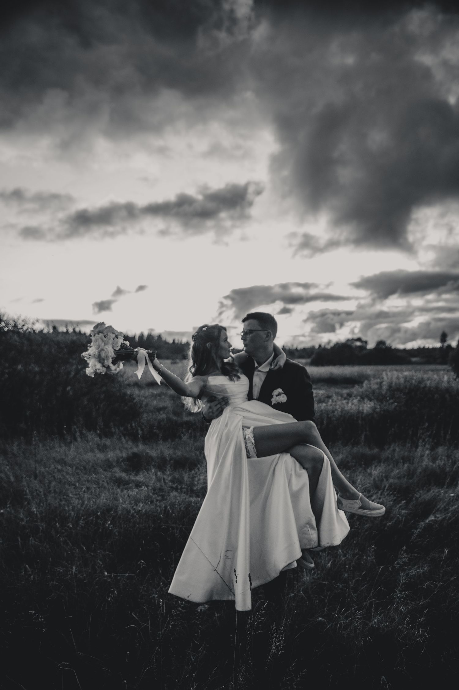 Dramatic black and white photo of couple in wedding attire posing in field at dusk.