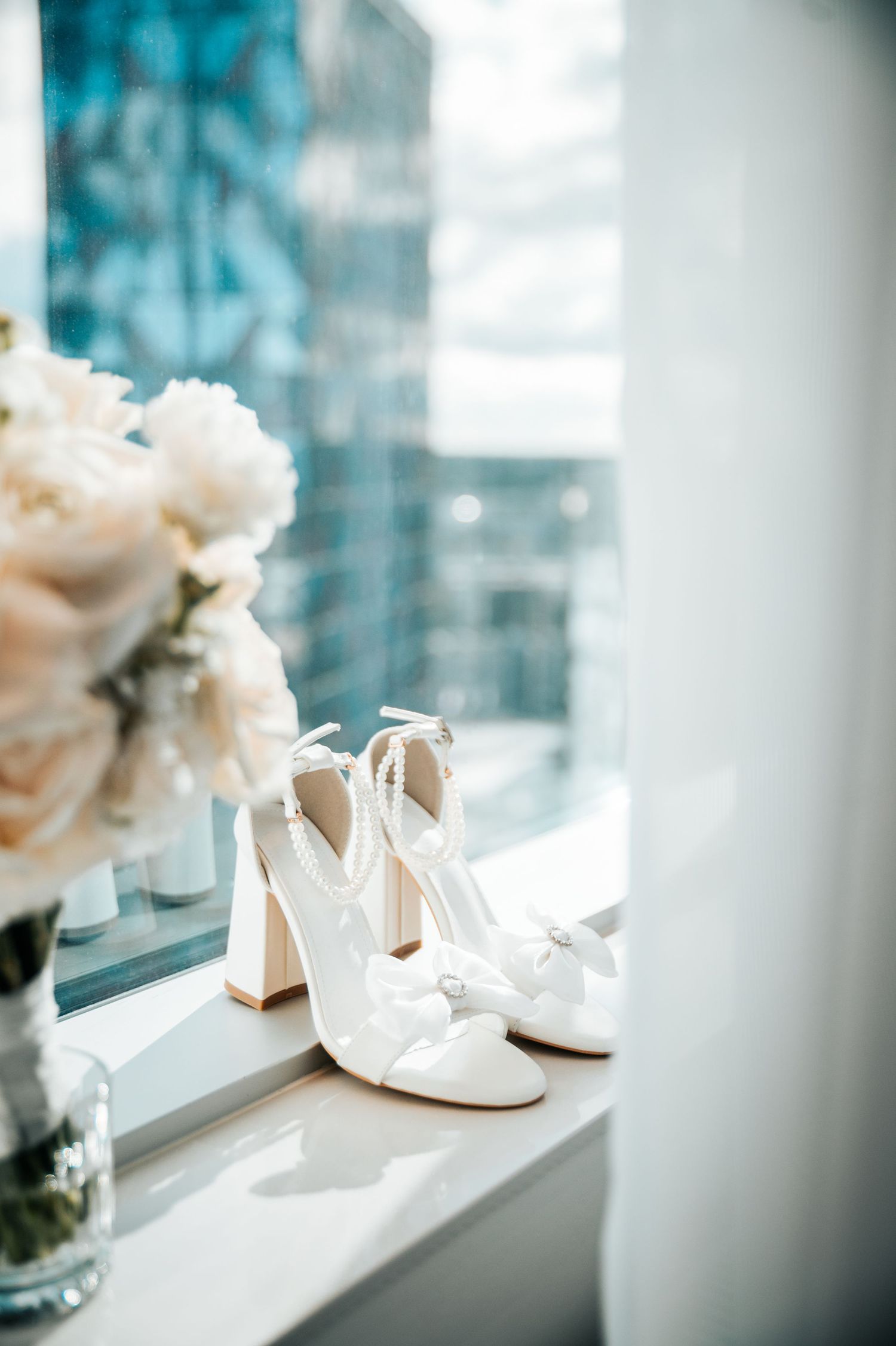 Elegant white wedding shoes with block heels and delicate floral arrangements displayed on a windowsill.