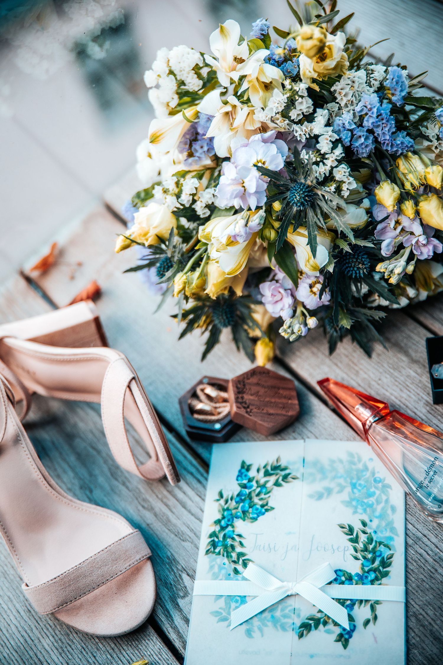 Wedding details with blue and yellow floral bouquet, invitation and blush pink shoes on wooden surface.