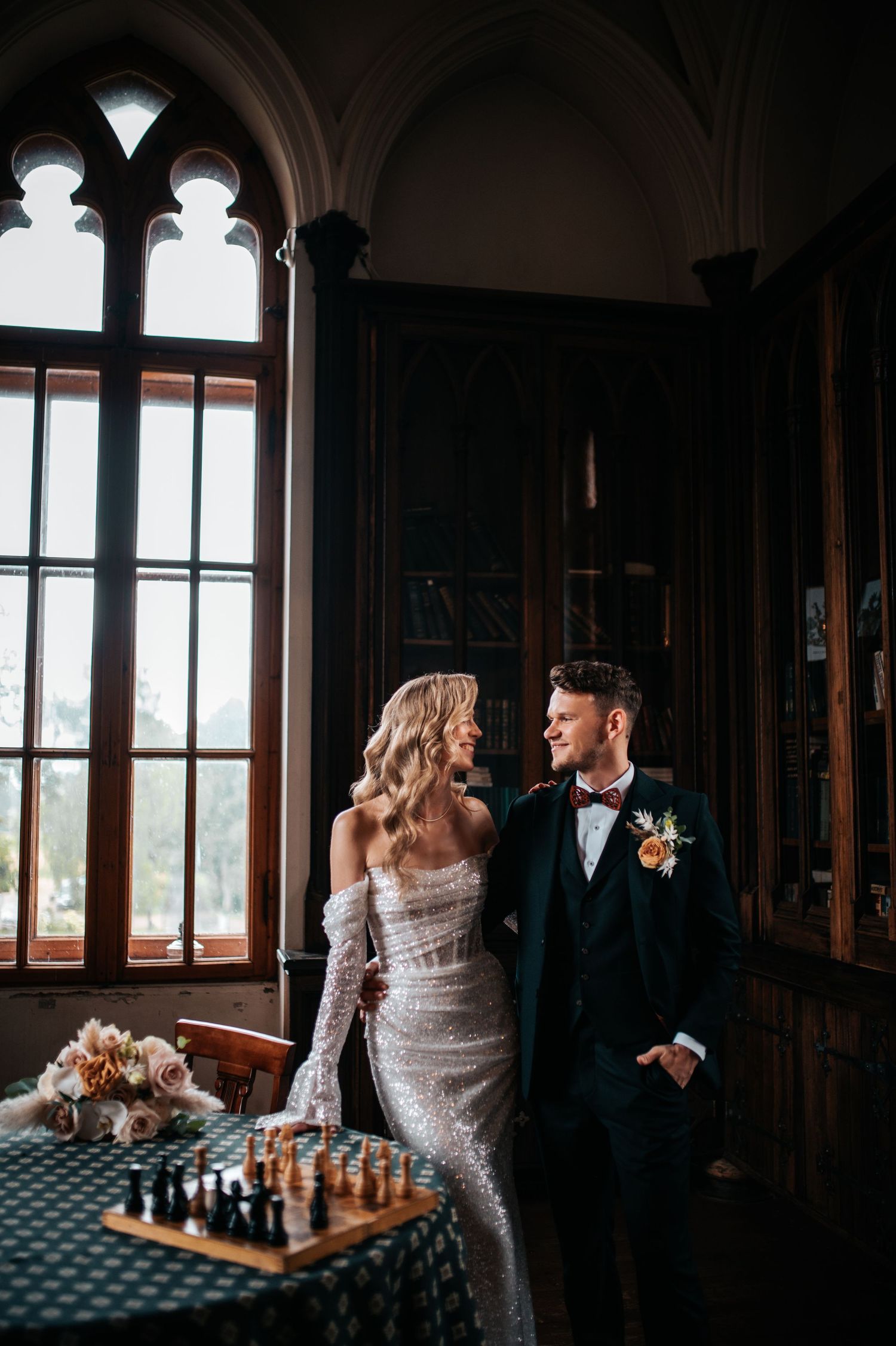 Elegant couple poses in a historic library with chess pieces and large windows creating a romantic atmosphere.