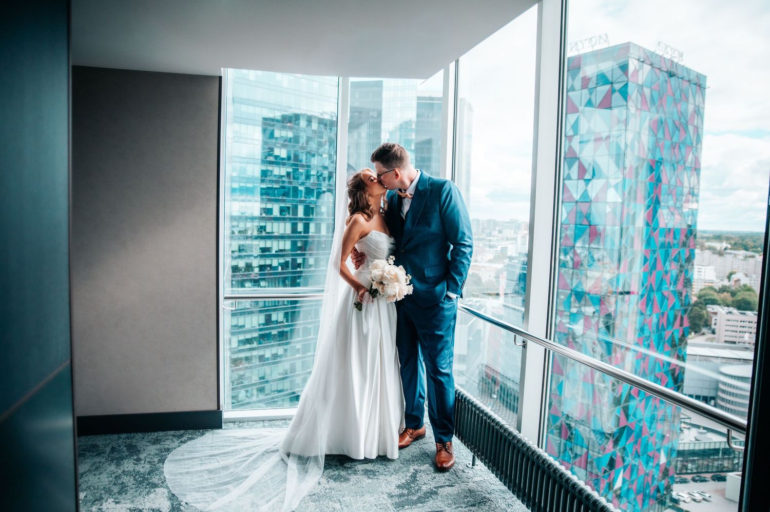 A romantic wedding couple shares intimate moments by large windows overlooking a city skyline.