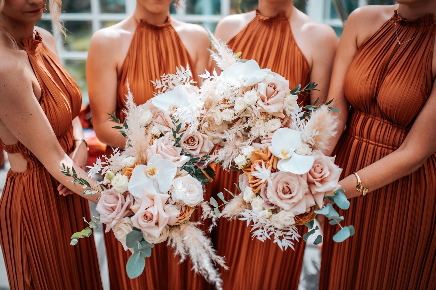 Bridesmaids in rust-colored dresses holding elegant white and blush floral bouquets.