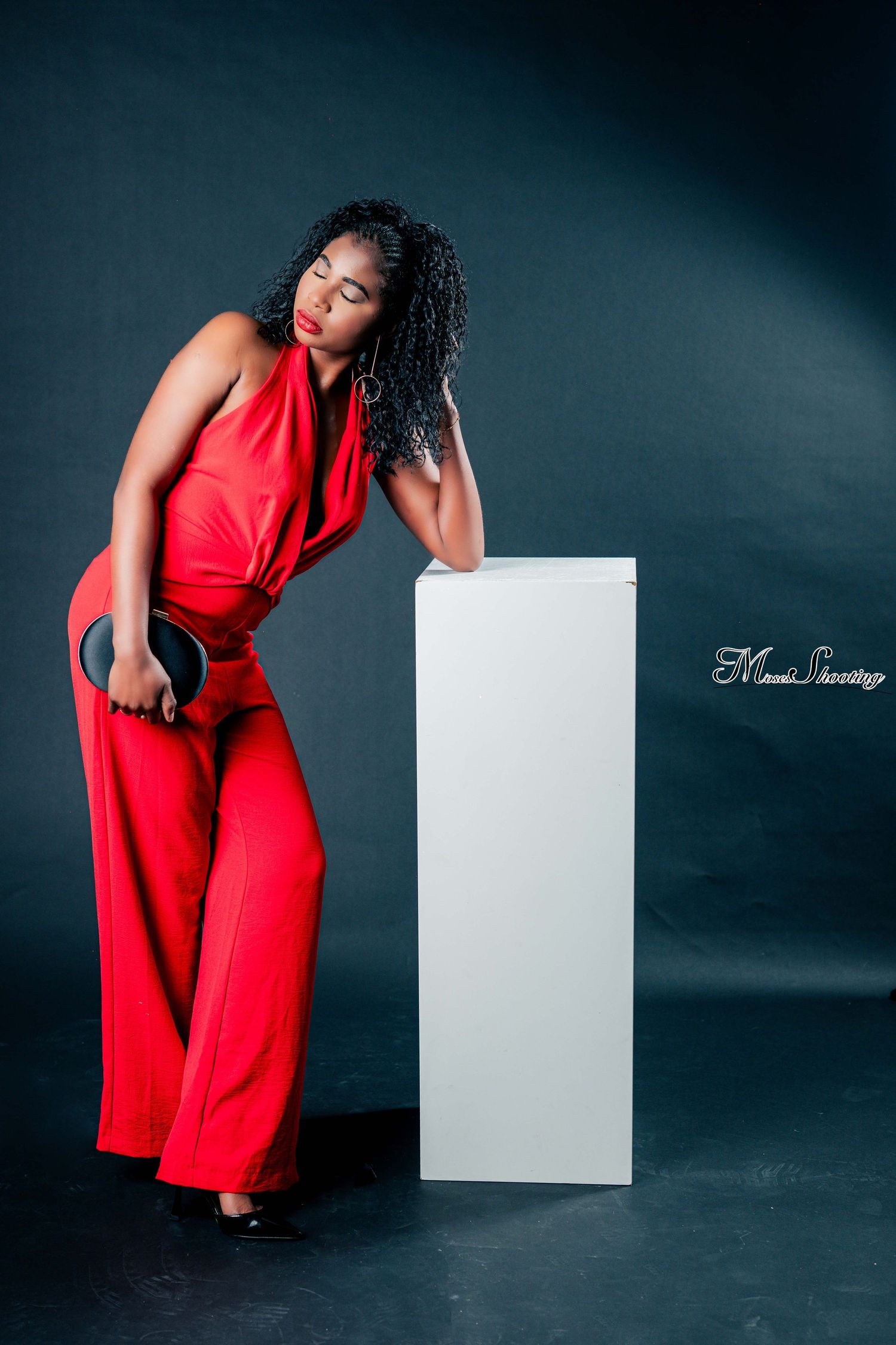 A model in a vibrant red jumpsuit poses elegantly against a dark backdrop with a white geometric prop.