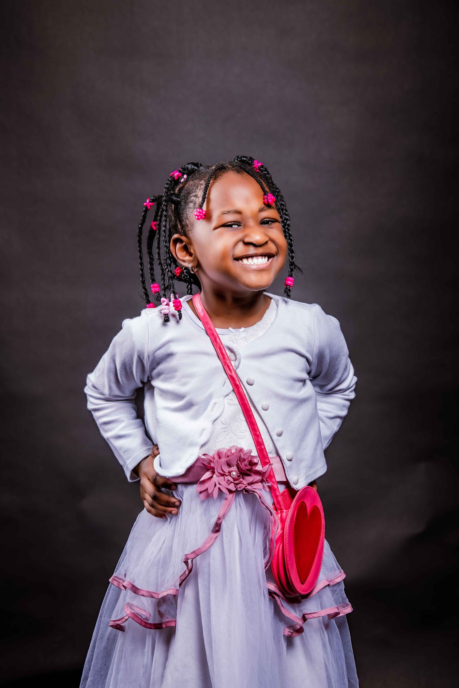Young child in a white dress with pink accents and braids smiles joyfully against dark background.
