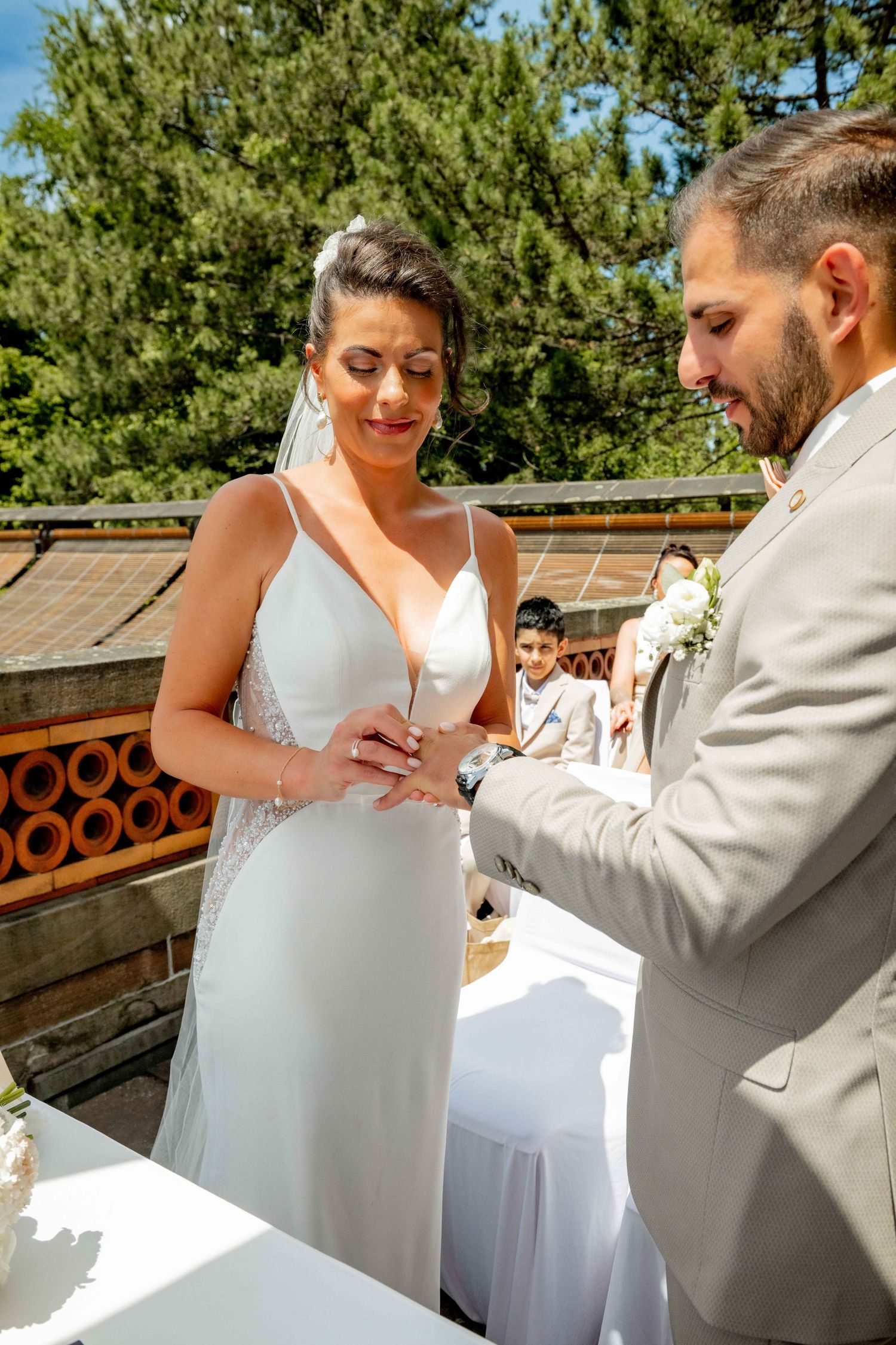 Bride and groom exchanging rings during outdoor wedding ceremony, bride in white gown smiling joyfully.