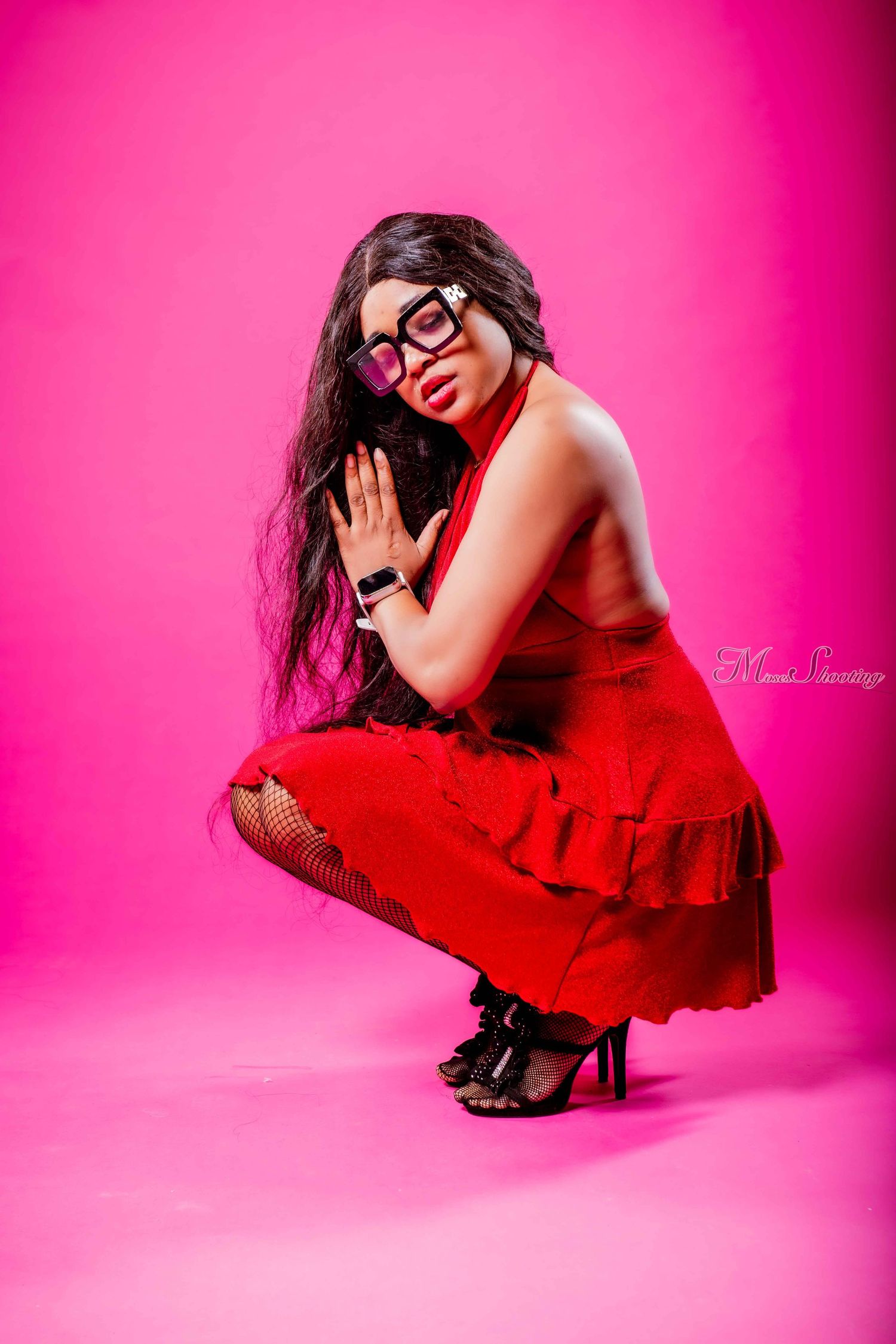 Fashion model in red ruffled dress and glasses poses dramatically against vibrant pink background.