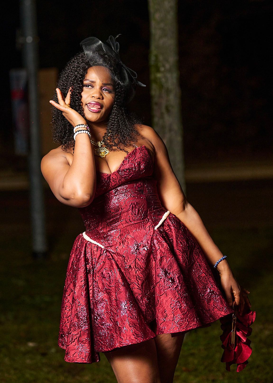 A person in a burgundy lace dress poses playfully outdoors during a photo session.
