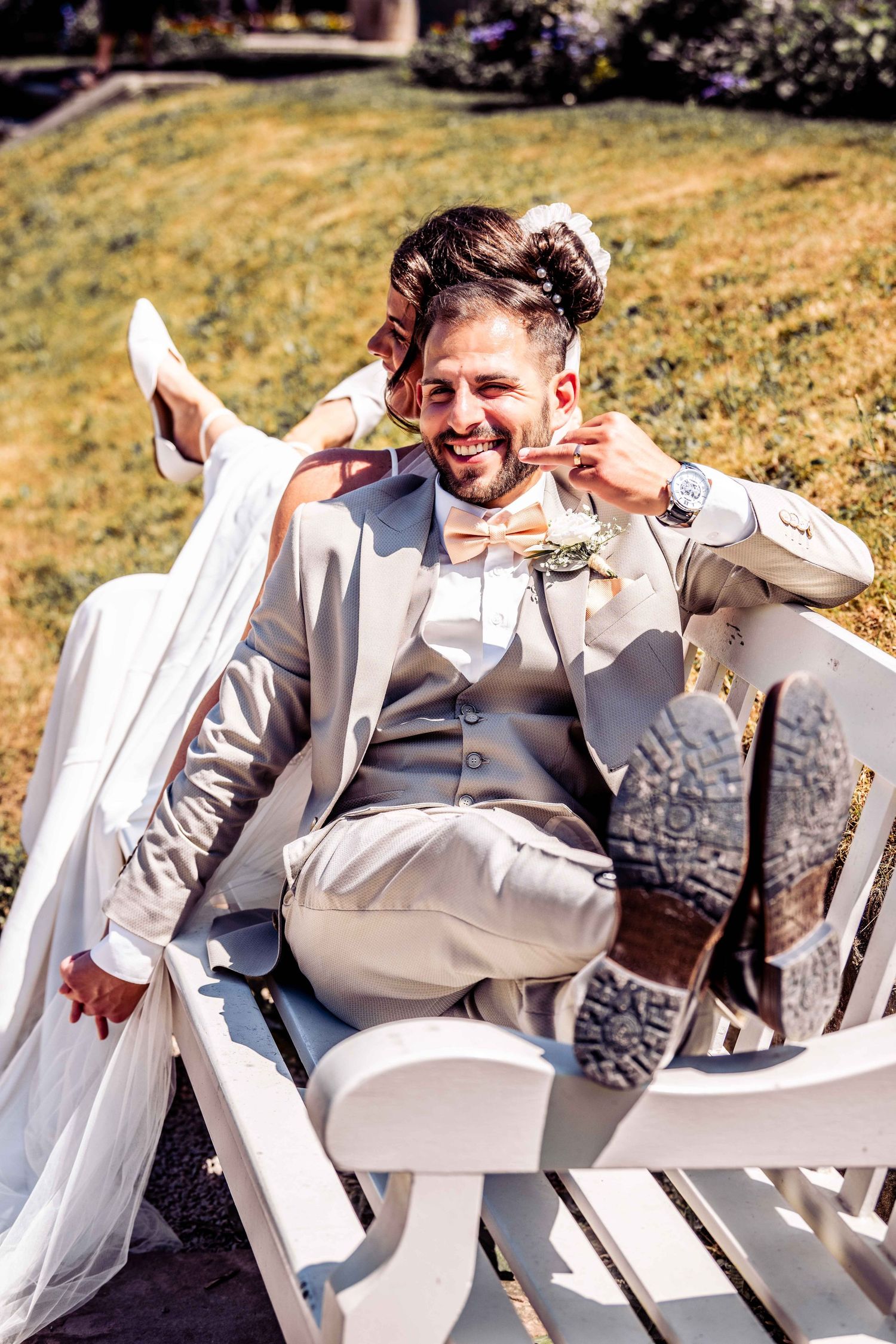 Smiling groom in gray suit relaxes on white bench while bride playfully rests legs on him outdoors.