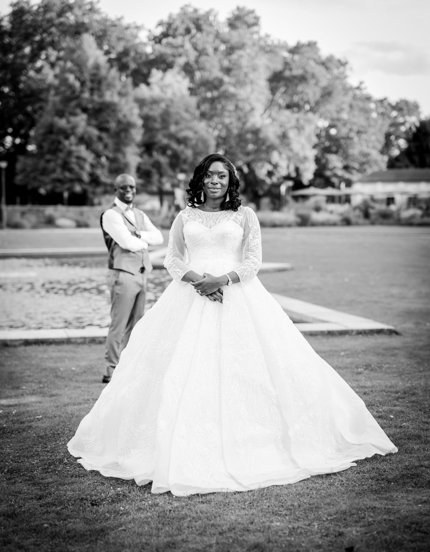 Black and white photo of a bride in a elegant ball gown standing on grass with a man in a suit behind her.