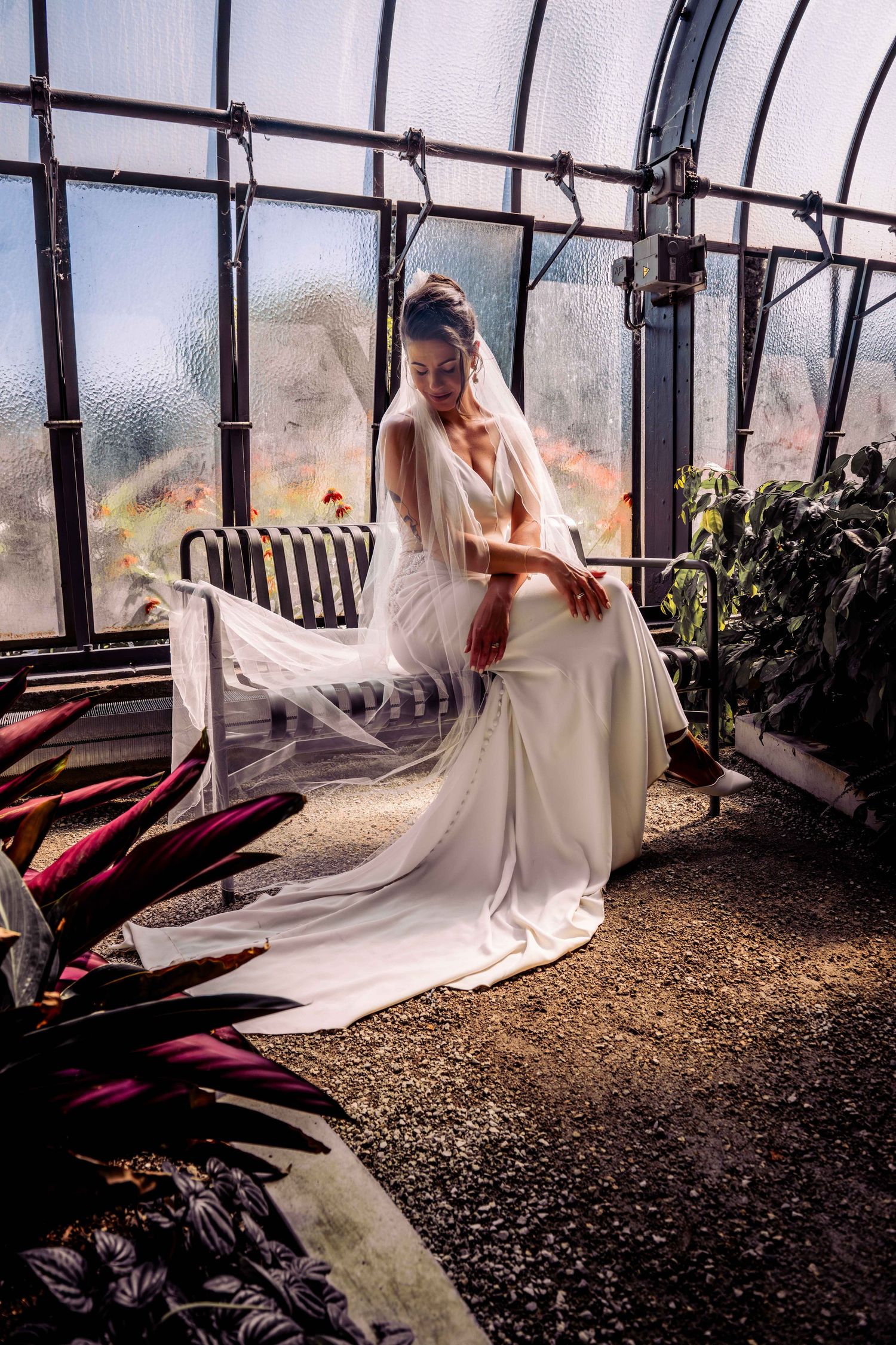 Elegant bride in a flowing white gown and veil seated in a sunlit greenhouse surrounded by lush plants.