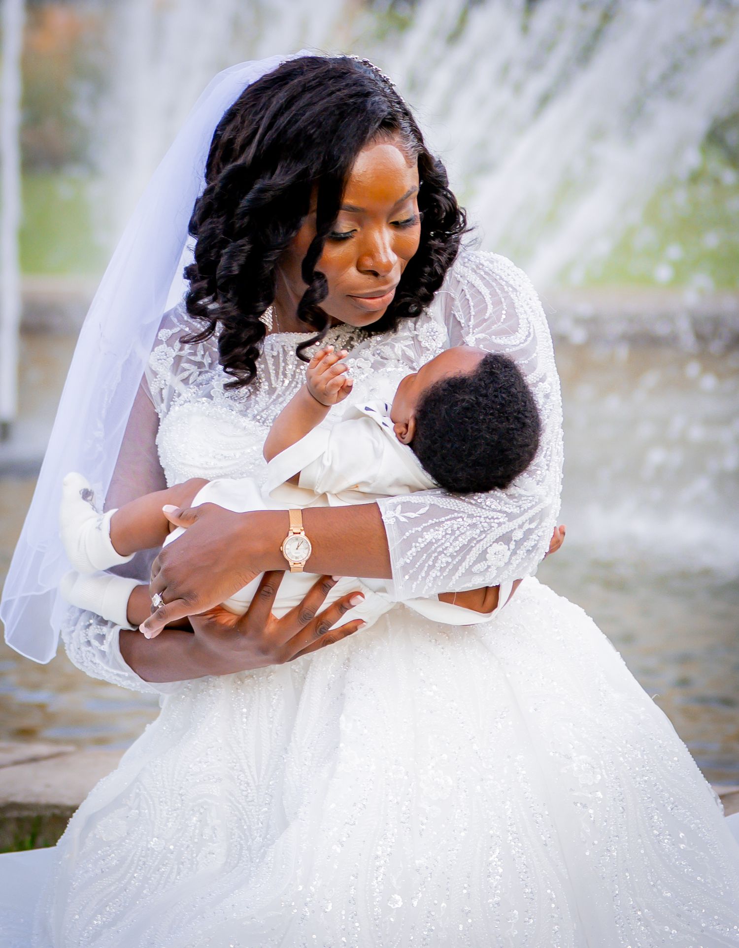 Bride in white lace gown and veil tenderly holds newborn baby near fountain on wedding day.