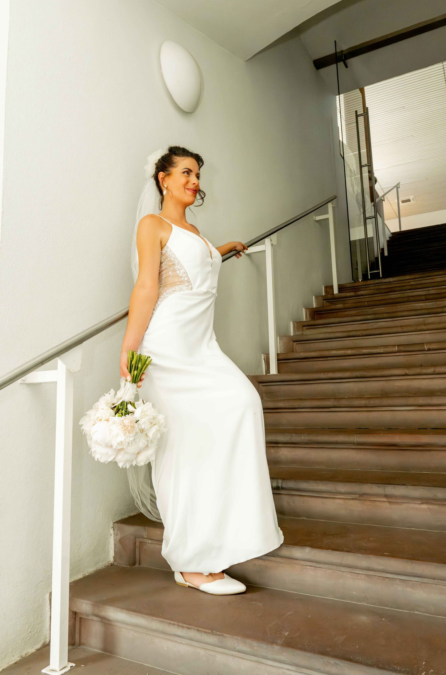 Elegant bride in a white sleeveless gown holds a peony bouquet while posing gracefully on a staircase.
