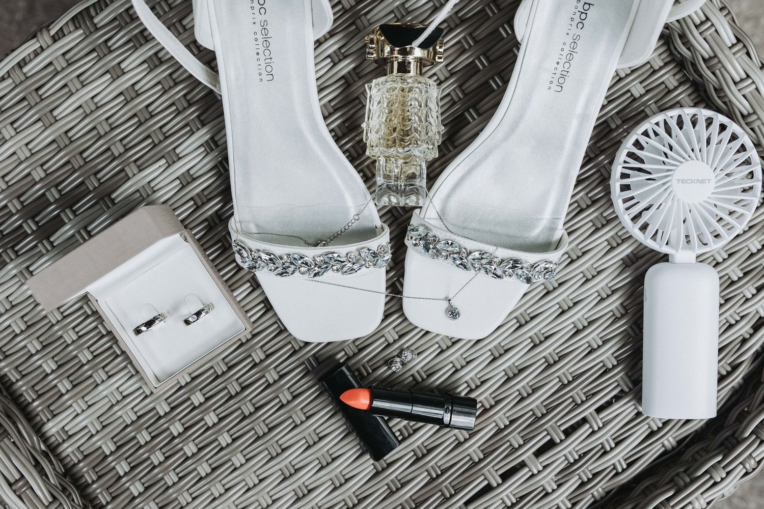 Wedding accessories including white shoes, perfume bottles, and jewelry arranged on a textured gray surface.