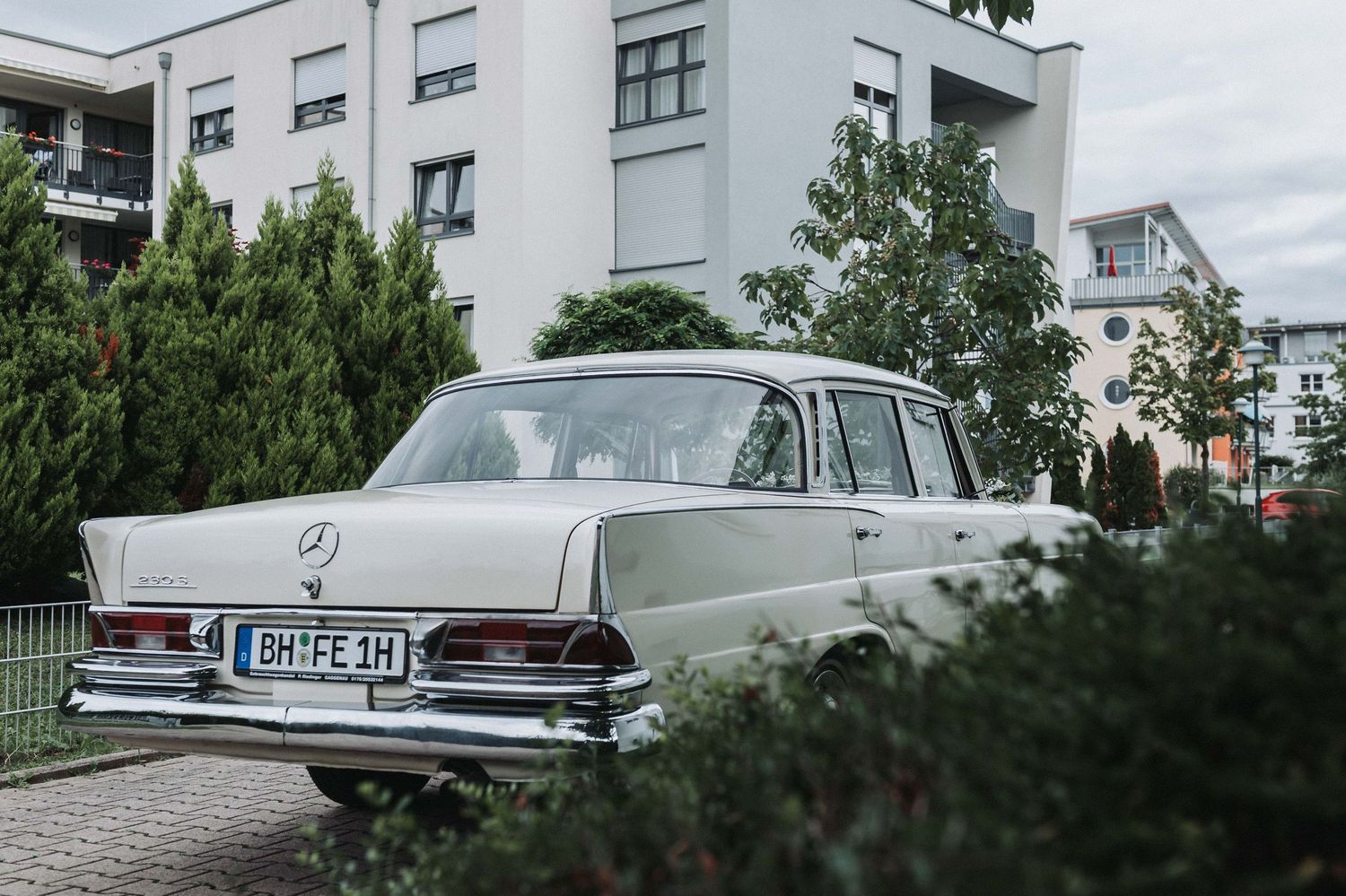 A classic white Mercedes-Benz sedan parked in front of a modern apartment building with trees and greenery.