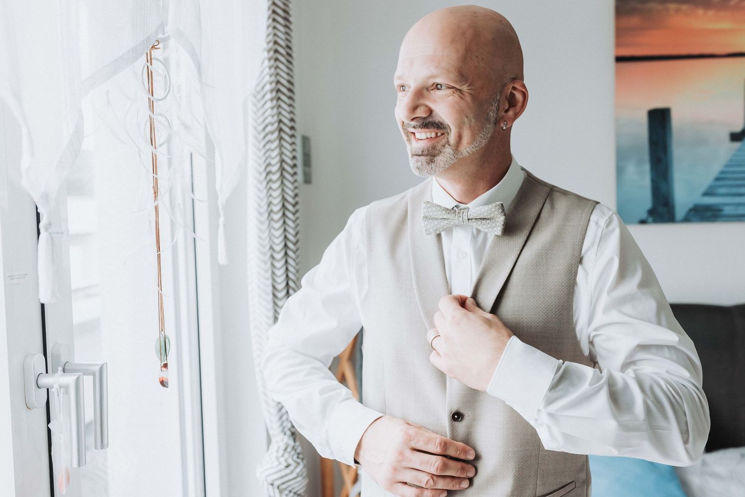 A groom in a light gray vest and white bow tie smiles joyfully while getting ready by a bright window.