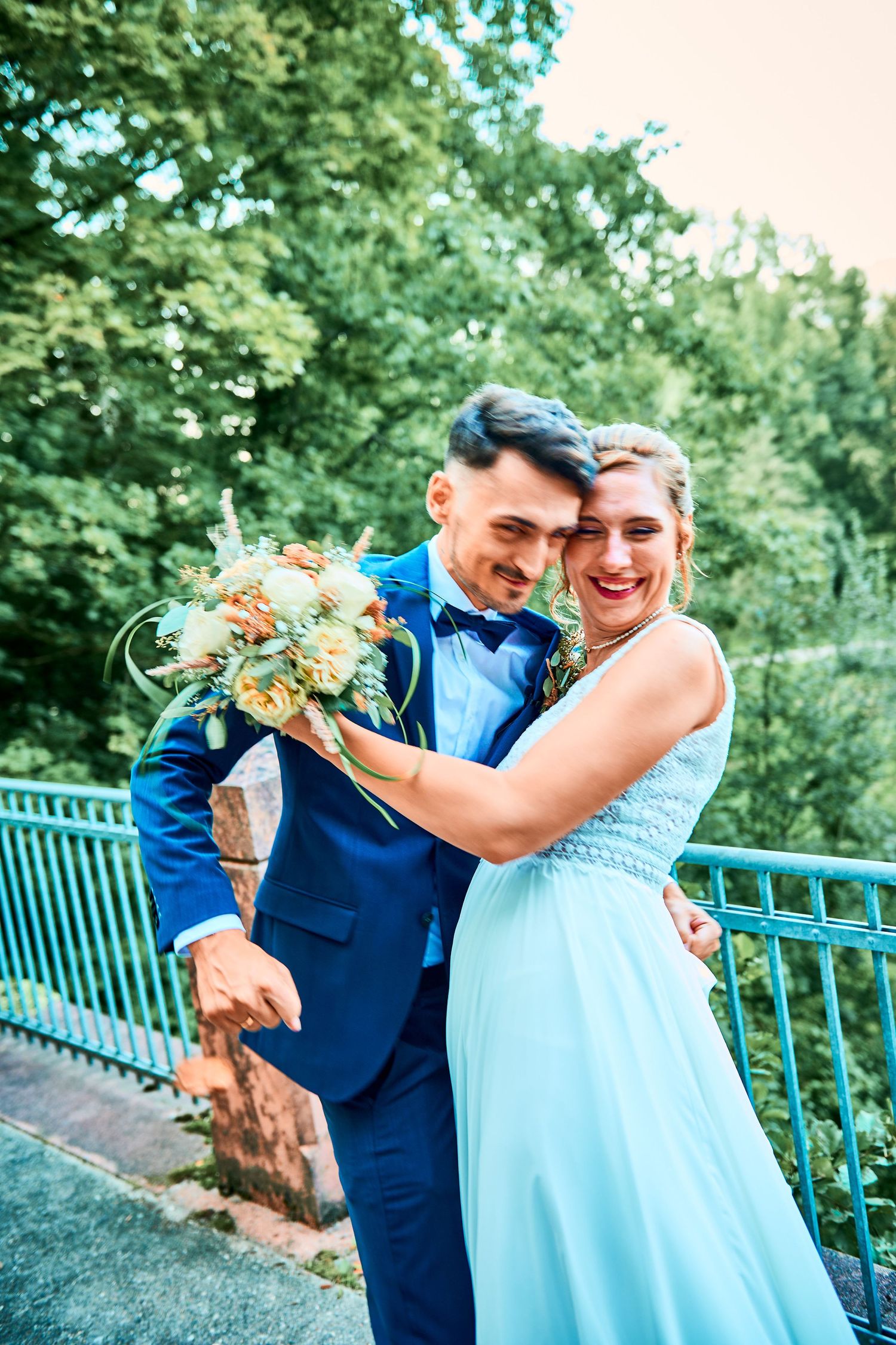 A couple shares a joyful moment together in a park setting with green trees and a blue railing in the background.