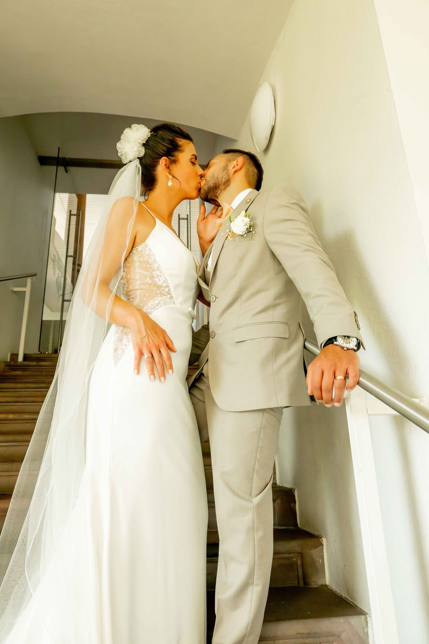 Bride and groom share a kiss on staircase, she in white lace gown with veil, he in beige suit.