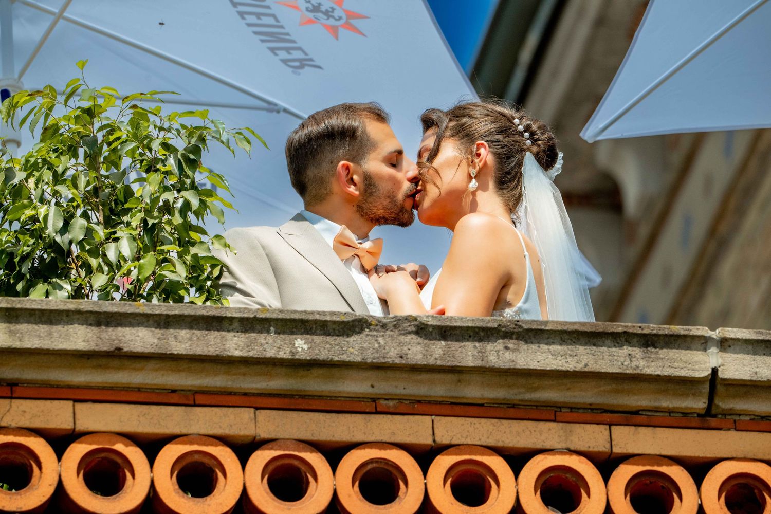 Bride and groom sharing a romantic kiss on a balcony, with lush greenery in the background on their wedding day.