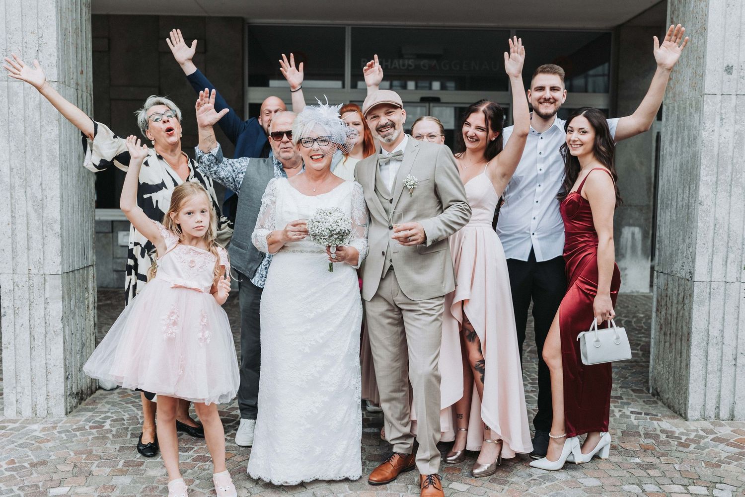 A joyful wedding party celebrates with raised arms and smiles in front of a rustic wooden door.
