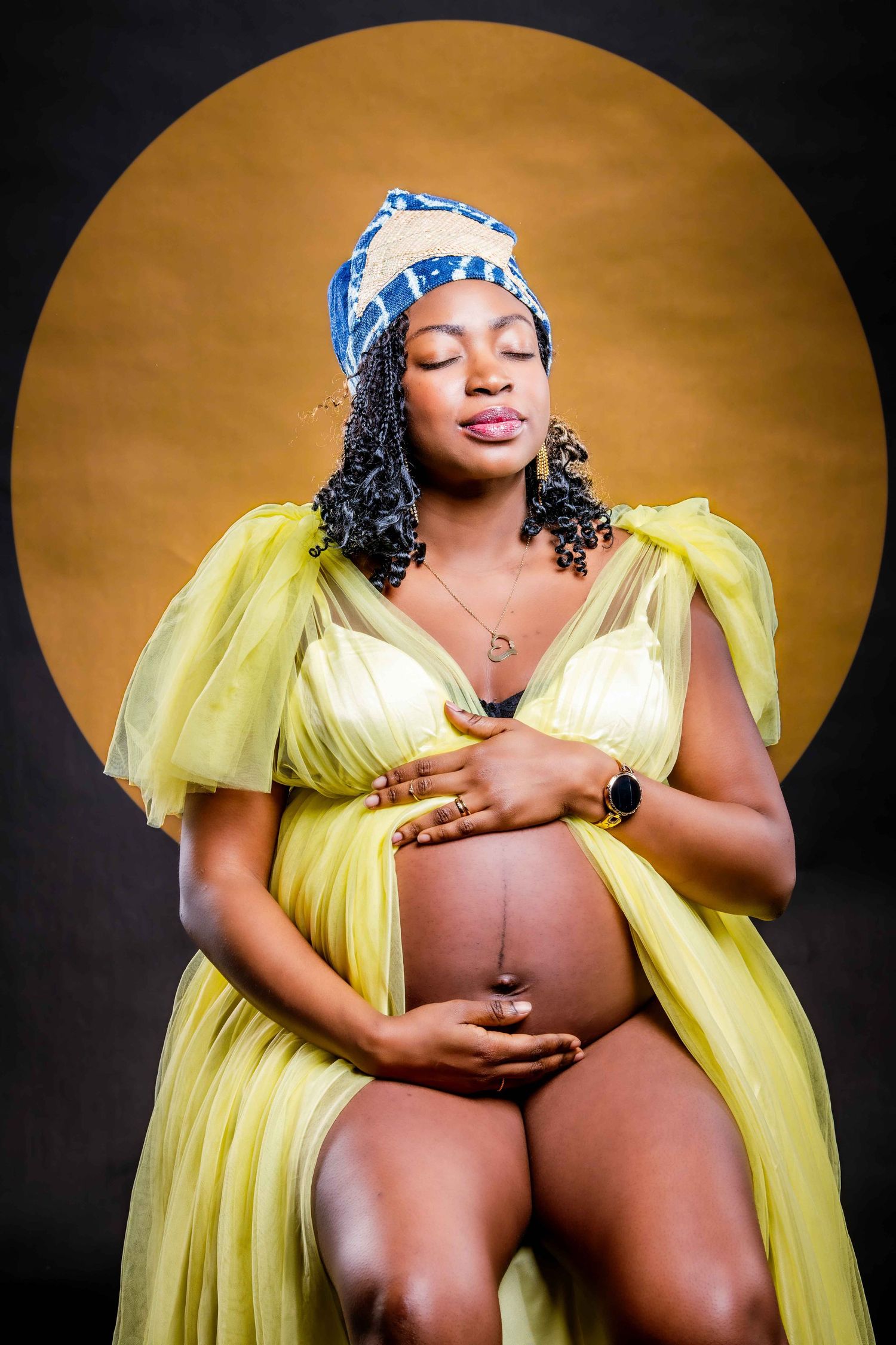 Maternity portrait of woman in yellow dress and blue headwrap against golden background.