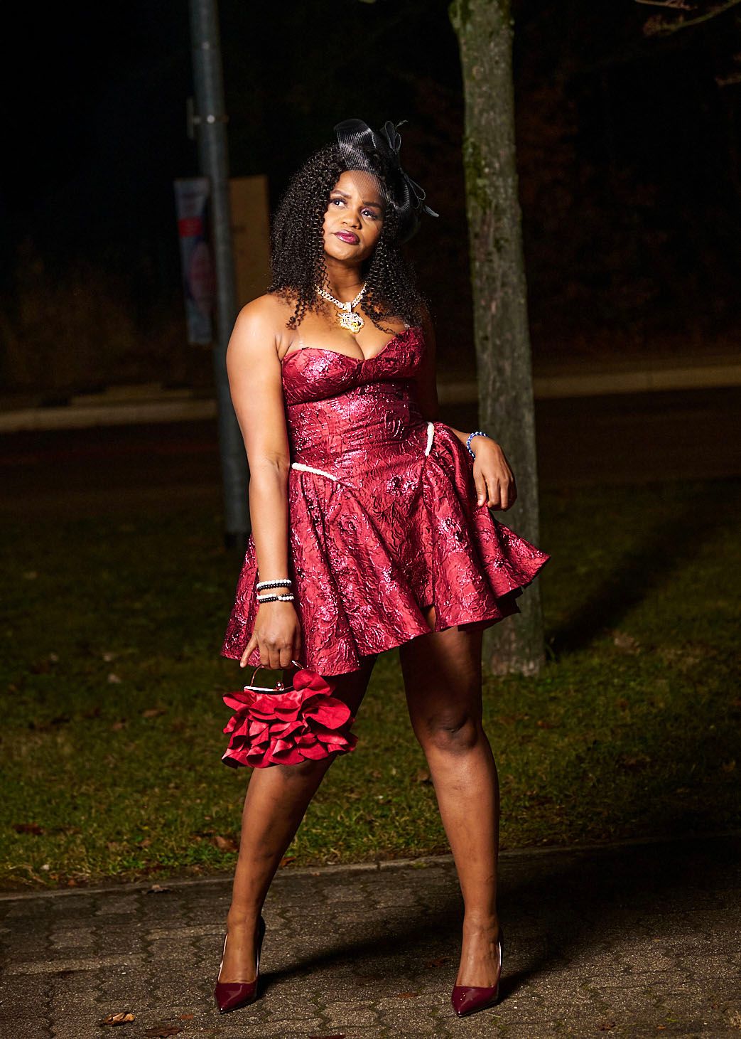A person in a burgundy velvet dress and matching heels poses outdoors at night against a dark background.